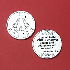 Op de afbeelding: Twee zilverkleurige metalen tokens met religieuze afbeeldingen. Een token toont een kruis met lichtstralen en een persoon met opgeheven armen. De andere token heeft de tekst "Vertrouw op de HEERE in al wat je doet, en je plannen zullen slagen." -Spreuken 16:3