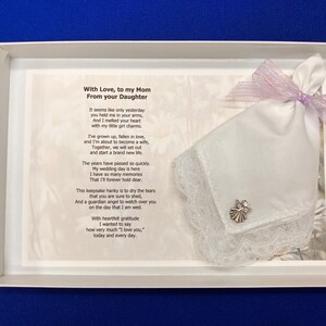 Puede incluir: Un pa&ntilde;uelo blanco con borde de encaje y un peque&ntilde;o dije de &aacute;ngel plateado, atado con una cinta morada clara, presentado en una caja blanca junto a un poema impreso titulado With Love, to my Mom From your Daughter, con un texto emotivo.