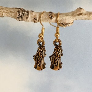 May include: A pair of gold-coloured earrings featuring a shoe design. The earrings are suspended from gold-coloured hooks and are displayed on a light-coloured branch against a soft blue background. The shoe pendants have intricate detailing.