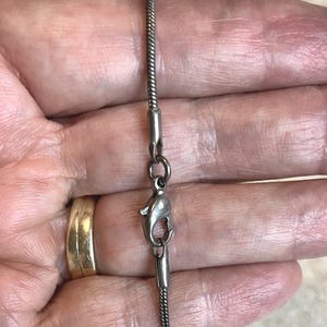 May include: Close-up of a silver-colored necklace with a lobster clasp. The chain is a thin, textured design. The clasp is attached to the chain with a small ring. A gold ring is visible in the background.
