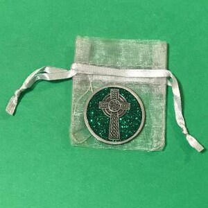 Puede incluir: Una moneda plateada con una cruz celta y un fondo verde brillante, presentada en una bolsa transparente plateada con cord&oacute;n. La moneda presenta intrincados dise&ntilde;os de nudos. La bolsa est&aacute; atada con una cinta de sat&eacute;n blanca, sobre una superficie verde.