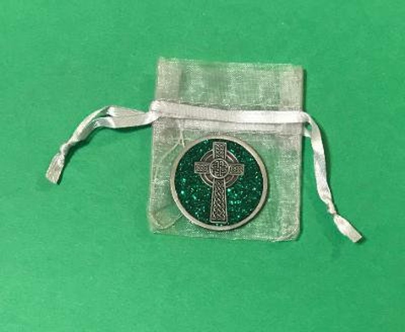 Puede incluir: Una moneda plateada con una cruz celta y un fondo verde brillante, presentada en una bolsa transparente plateada con cord&oacute;n. La moneda presenta intrincados dise&ntilde;os de nudos. La bolsa est&aacute; atada con una cinta de sat&eacute;n blanca, sobre una superficie verde.