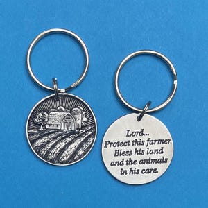 May include: Two silver keychains. One keychain features a detailed engraving of a farm scene with a barn, silo, and fields. The other keychain has the text: "Lord... Protect this farmer. Bless his land and the animals in his care."
