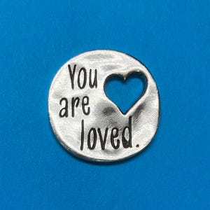 Puede incluir: Un amuleto ovalado de plata con las palabras "You are loved" y un corazón recortado. El corazón está relleno con un fondo azul.