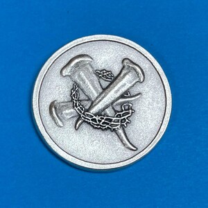 Pu&ograve; includere: Una moneta rotonda color argento con un design dettagliato di due chiodi incrociati e una corona di spine. La moneta ha una superficie strutturata e un bordo rialzato, creando un effetto tridimensionale.