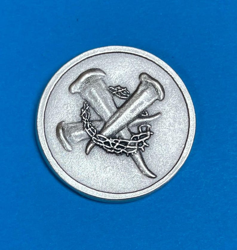 Pu&ograve; includere: Una moneta rotonda color argento con un design dettagliato di due chiodi incrociati e una corona di spine. La moneta ha una superficie strutturata e un bordo rialzato, creando un effetto tridimensionale.