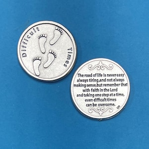 Peut inclure: Deux pièces de monnaie argentées. L'une porte l'inscription "Difficult Times" sur le bord et quatre empreintes de pas au centre. L'autre pièce porte une citation inspirante sur la foi et le dépassement des difficultés.