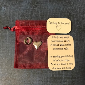 May include: A small red pouch with a gold charm of a heart and a letter O. The pouch has a card inside with the text "This hug is for you" and a poem about hugs.