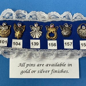 May include: A display of six angel-themed pins in gold and silver finishes, each with a rhinestone accent. The pins are arranged on a blue fabric, framed by white lace. The text reads: "All pins are available in gold or silver finishes."