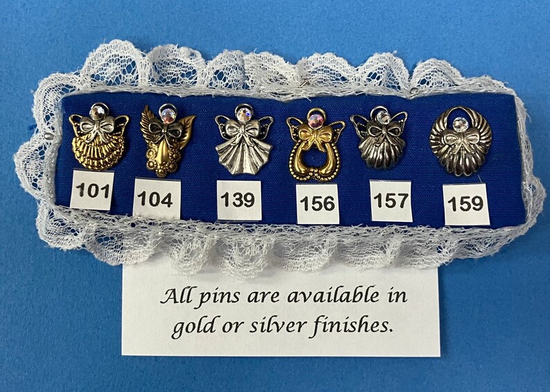 May include: A display of six angel-themed pins in gold and silver finishes, each with a rhinestone accent. The pins are arranged on a blue fabric, framed by white lace. The text reads: "All pins are available in gold or silver finishes."