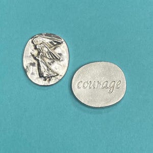Peut inclure: Deux pendentifs argent&eacute;s. L'un est de forme ovale avec un relief d'une figure ail&eacute;e. L'autre est rond avec le mot "courage" en cursive. Les pendentifs sont sur un fond turquoise.
