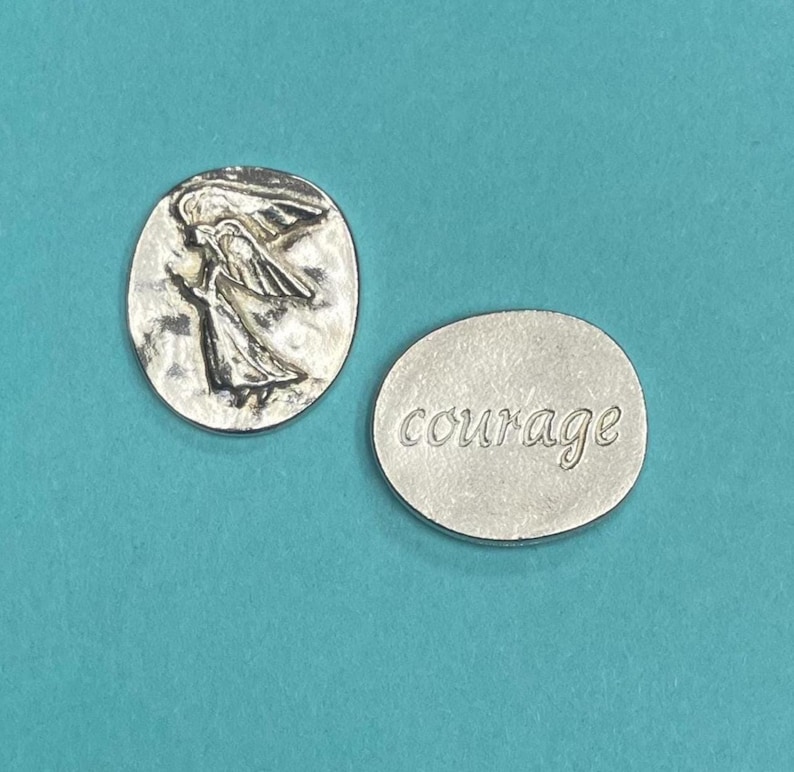 Peut inclure: Deux pendentifs argent&eacute;s. L'un est de forme ovale avec un relief d'une figure ail&eacute;e. L'autre est rond avec le mot "courage" en cursive. Les pendentifs sont sur un fond turquoise.
