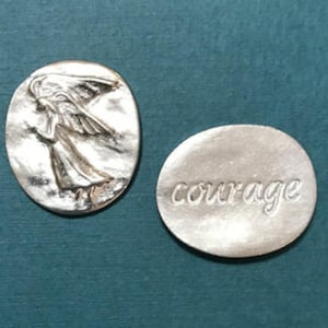Puede incluir: Dos fichas de metal plateadas. Una ficha presenta una imagen en relieve de un ángel con alas. La otra ficha tiene la palabra "coraje" en relieve.