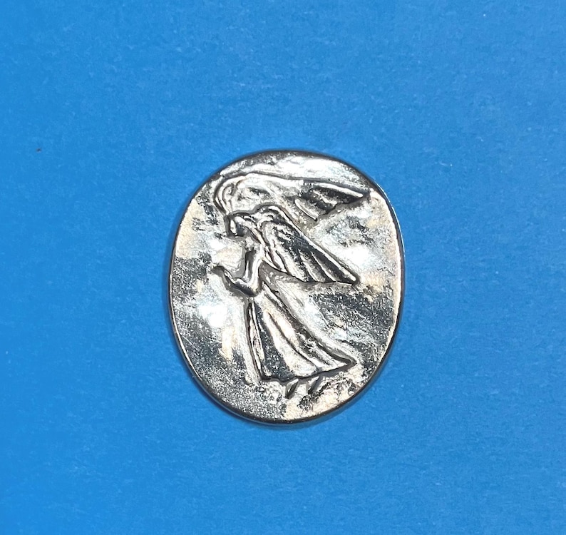 Peut inclure: Un pendentif ovale argent&eacute; repr&eacute;sentant un ange en relief avec des ailes d&eacute;ploy&eacute;es et des robes fluides. L'ange est repr&eacute;sent&eacute; dans une pose dynamique, sur un fond textur&eacute;. Le pendentif est centr&eacute; sur un fond bleu uni.