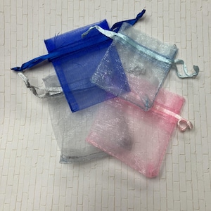 Könnte beinhalten: Vier kleine, transparente Kordelzugbeutel in Blau, Silber und Rosa. Die Beutel bestehen aus einem leichten, netzartigen Material. Jeder Beutel hat einen passenden Kordelzugverschluss aus Satinband. Die Beutel sind auf einer weißen, ziegelgemusterten Oberfläche angeordnet.