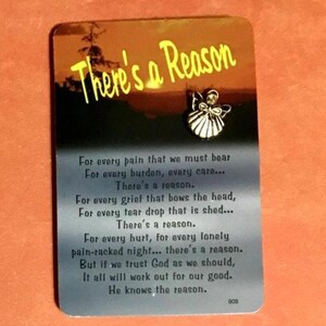 May include: A rectangular card with the text "There's a Reason" in yellow script. The card features a small silver angel pin and a poem about finding reason in pain and grief. The background is a sunset scene.