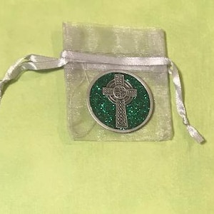 Puede incluir: Una moneda plateada con un dise&ntilde;o de cruz celta sobre un fondo verde brillante. La moneda est&aacute; dentro de una peque&ntilde;a bolsa transparente atada con plata, sobre una superficie verde claro. El di&aacute;metro de la moneda es de aproximadamente 3,8 cm.