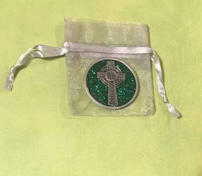 Puede incluir: Una moneda plateada con un dise&ntilde;o de cruz celta sobre un fondo verde brillante. La moneda est&aacute; dentro de una peque&ntilde;a bolsa transparente atada con plata, sobre una superficie verde claro. El di&aacute;metro de la moneda es de aproximadamente 3,8 cm.