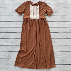 May include: A brown ribbed-knit dress with short sleeves and a round neckline. The bodice features a vertical panel of white lace. The dress has a gathered waist and a flowing, floor-length skirt. The dress is displayed on a light wood-grain surface.