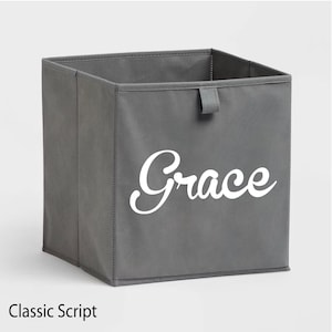 May include: A gray storage bin with the name "Grace" in white script. The bin has a rectangular shape with a small handle on the top edge. The text "Classic Script" is at the bottom.