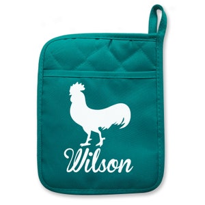 May include: Teal pot holder with a white rooster silhouette and the name "Wilson" in cursive. The pot holder has a quilted pattern and a small pocket. It also has a loop for hanging.