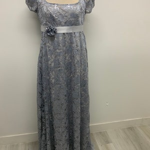 Könnte beinhalten: Ein silbernes Pailletten-Spitzen-Maxikleid mit einem weißen Satinschleifenband und einem Blumenakzent. Das Kleid hat kurze Ärmel und einen quadratischen Ausschnitt.