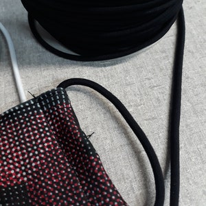 Puede incluir: Primer plano de un cordón elástico negro enrollado en una bobina, con una máscara de tela con estampado rojo y blanco en primer plano.