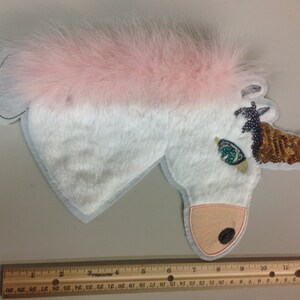May include: A white unicorn applique with a pink fur mane and a gold sequined horn. The unicorn has a green eye and a brown nose.