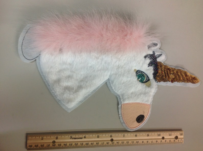 May include: A white unicorn applique with a pink fur mane and a gold sequined horn. The unicorn has a green eye and a brown nose.