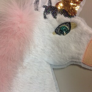 May include: A white unicorn applique with pink fur, a gold sequin horn, and a green sequin eye. The unicorn is embroidered with black thread.