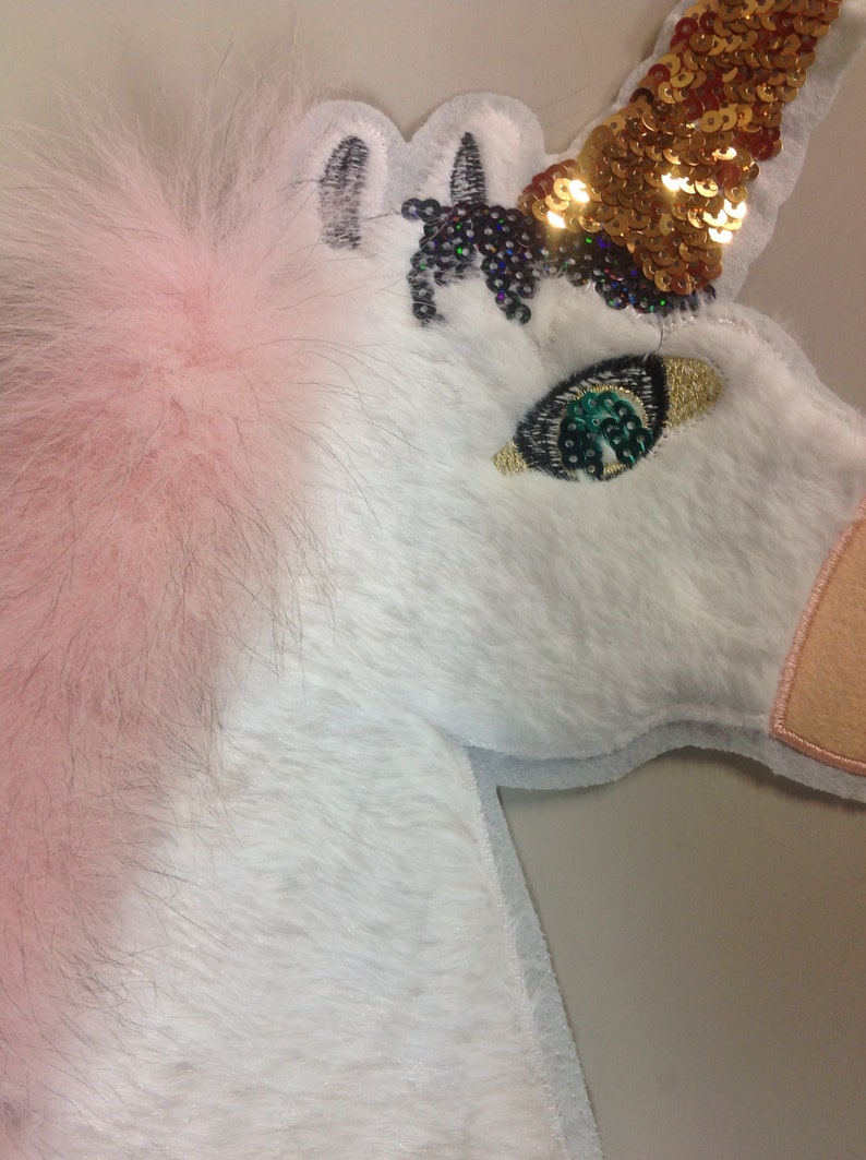 May include: A white unicorn applique with pink fur, a gold sequin horn, and a green sequin eye. The unicorn is embroidered with black thread.