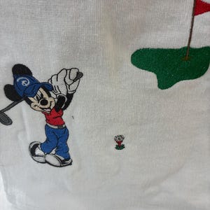 K&ouml;nnte beinhalten: Ein wei&szlig;es Handtuch mit einer Stickerei einer Zeichentrickmaus, die eine blaue Kappe, ein rotes Oberteil und eine blaue Hose tr&auml;gt und einen Golfschl&auml;ger schwingt. Das Motiv zeigt zudem einen Golfball auf einem roten Tee und ein gr&uuml;nes Putting-Green.