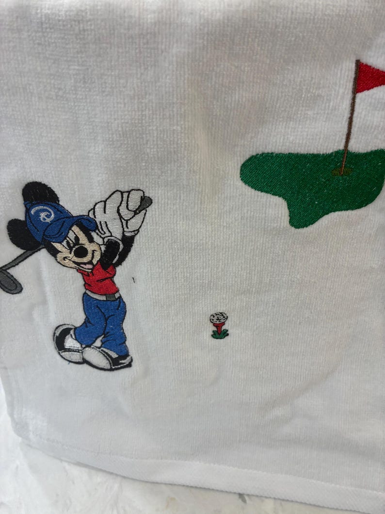 K&ouml;nnte beinhalten: Ein wei&szlig;es Handtuch mit einer Stickerei einer Zeichentrickmaus, die eine blaue Kappe, ein rotes Oberteil und eine blaue Hose tr&auml;gt und einen Golfschl&auml;ger schwingt. Das Motiv zeigt zudem einen Golfball auf einem roten Tee und ein gr&uuml;nes Putting-Green.