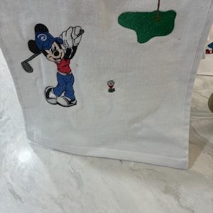 K&ouml;nnte beinhalten: Ein wei&szlig;es Handtuch mit einer Stickerei einer Zeichentrickmaus, die eine blaue Kappe, ein rotes Oberteil und eine blaue Hose tr&auml;gt und einen Golfschl&auml;ger schwingt. Das Motiv zeigt zudem einen Golfball auf einem roten Tee und ein gr&uuml;nes Putting-Green.