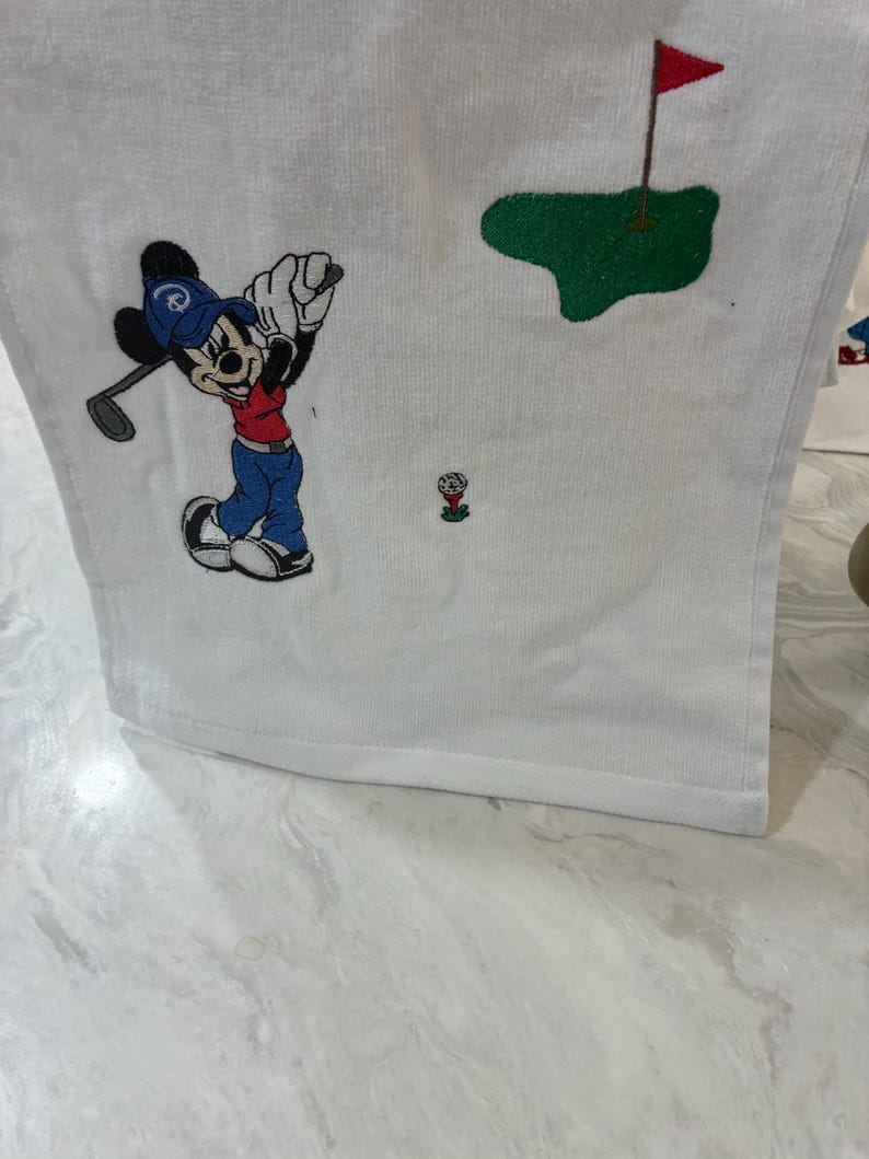 K&ouml;nnte beinhalten: Ein wei&szlig;es Handtuch mit einer Stickerei einer Zeichentrickmaus, die eine blaue Kappe, ein rotes Oberteil und eine blaue Hose tr&auml;gt und einen Golfschl&auml;ger schwingt. Das Motiv zeigt zudem einen Golfball auf einem roten Tee und ein gr&uuml;nes Putting-Green.