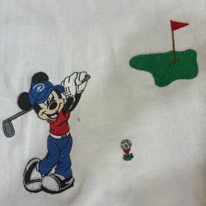 K&ouml;nnte beinhalten: Ein wei&szlig;er Stoff mit einer Stickerei von Mickey Mouse, der eine blaue Kappe, ein rotes Oberteil und eine blaue Hose tr&auml;gt und einen Golfschl&auml;ger schwingt. Ein kleiner Golfball auf einem roten Tee und ein gr&uuml;nes Putting Green mit roter Fahne.