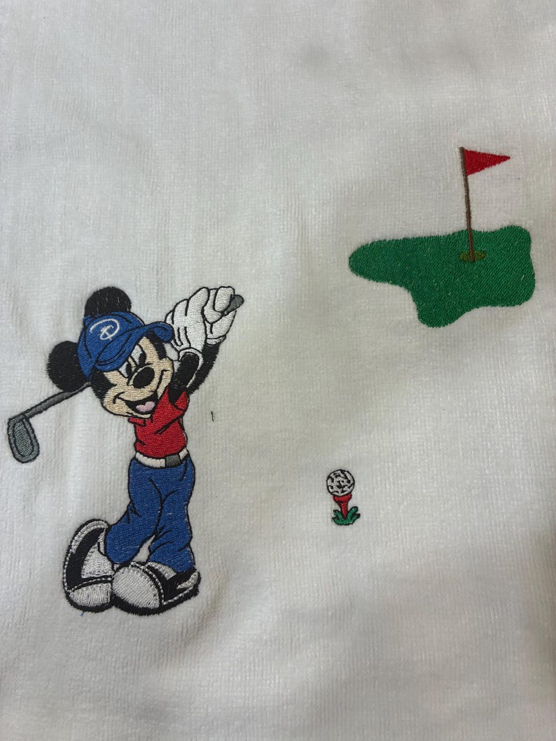 K&ouml;nnte beinhalten: Ein wei&szlig;er Stoff mit einer Stickerei von Mickey Mouse, der eine blaue Kappe, ein rotes Oberteil und eine blaue Hose tr&auml;gt und einen Golfschl&auml;ger schwingt. Ein kleiner Golfball auf einem roten Tee und ein gr&uuml;nes Putting Green mit roter Fahne.