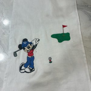 K&ouml;nnte beinhalten: Ein wei&szlig;es, rechteckiges Golftuch mit einer Stickerei von Mickey Mouse, die eine blaue M&uuml;tze, ein rotes Oberteil und eine blaue Hose tr&auml;gt und einen Golfschl&auml;ger schwingt. Das Design zeigt ein gr&uuml;nes Putting-Green mit roter Fahne und einen Golfball.