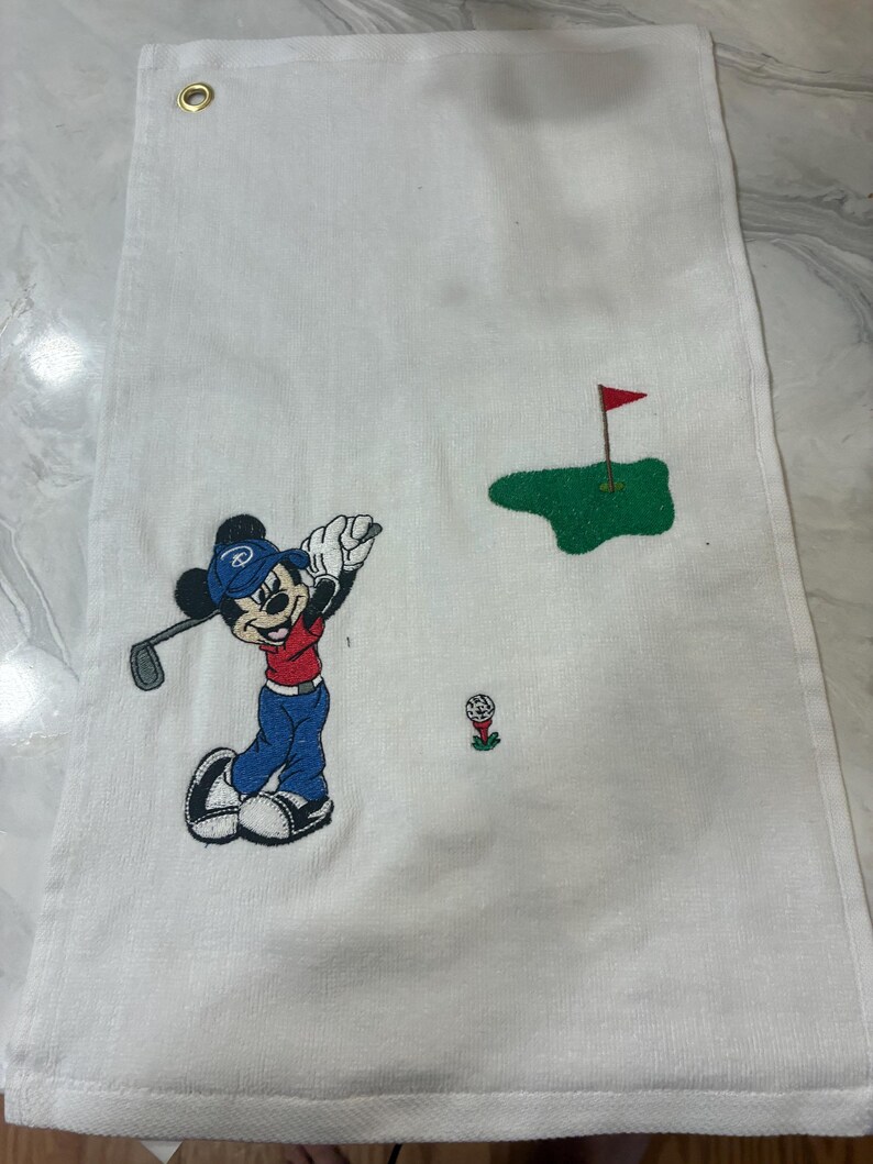 K&ouml;nnte beinhalten: Ein wei&szlig;es, rechteckiges Golftuch mit einer Stickerei von Mickey Mouse, die eine blaue M&uuml;tze, ein rotes Oberteil und eine blaue Hose tr&auml;gt und einen Golfschl&auml;ger schwingt. Das Design zeigt ein gr&uuml;nes Putting-Green mit roter Fahne und einen Golfball.