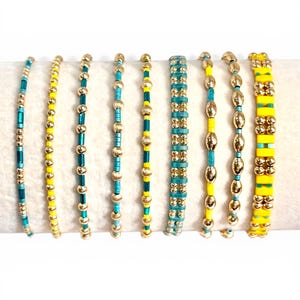 May include: A collection of beaded bracelets in various designs. The bracelets feature a combination of gold, turquoise, and yellow beads. Some bracelets have a linear design, while others have a more clustered appearance. The bracelets are displayed on a white surface.