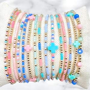 May include: A collection of beaded bracelets in pastel colors, including pink, blue, and turquoise, with gold-colored beads. Some bracelets feature cross-shaped charms. The bracelets are displayed on a white surface.