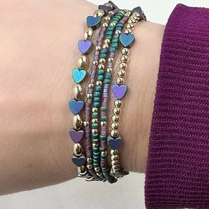 May include: A set of five bracelets with iridescent heart-shaped beads and gold beads. The bracelets are stacked on a person's wrist.