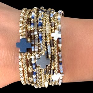 May include: A stack of beaded bracelets in gold, white, blue, and gray. The bracelets feature small beads and cross-shaped charms. The bracelets are worn on a wrist against a black background.