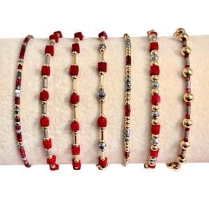 May include: A collection of seven beaded bracelets. The bracelets feature a variety of designs, including red square beads, gold beads, and silver beads. Some bracelets have gold or silver bars between the beads. The bracelets are displayed on a cream-colored surface.