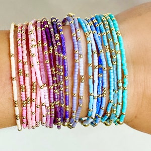 May include: A collection of beaded bracelets in various colors, including white, pink, purple, and blue. Each bracelet features small, colorful beads interspersed with gold-colored beads. The bracelets are stacked on a wrist, showcasing the range of colors and designs.