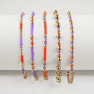 May include: Five beaded bracelets in a row. The bracelets feature a mix of small, colorful beads in orange, purple, and gold. The bracelets are made of small, round beads and larger, gold-colored beads.