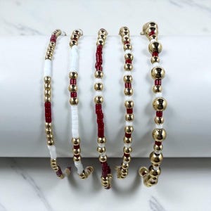 May include: Five beaded bracelets. Each bracelet features a combination of gold-colored beads, white beads, and red beads. The bracelets are displayed on a white, curved surface against a white background. The beads are strung together to create a flexible design.