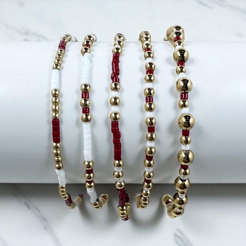 May include: Five beaded bracelets. Each bracelet features a combination of gold-colored beads, white beads, and red beads. The bracelets are displayed on a white, curved surface against a white background. The beads are strung together to create a flexible design.