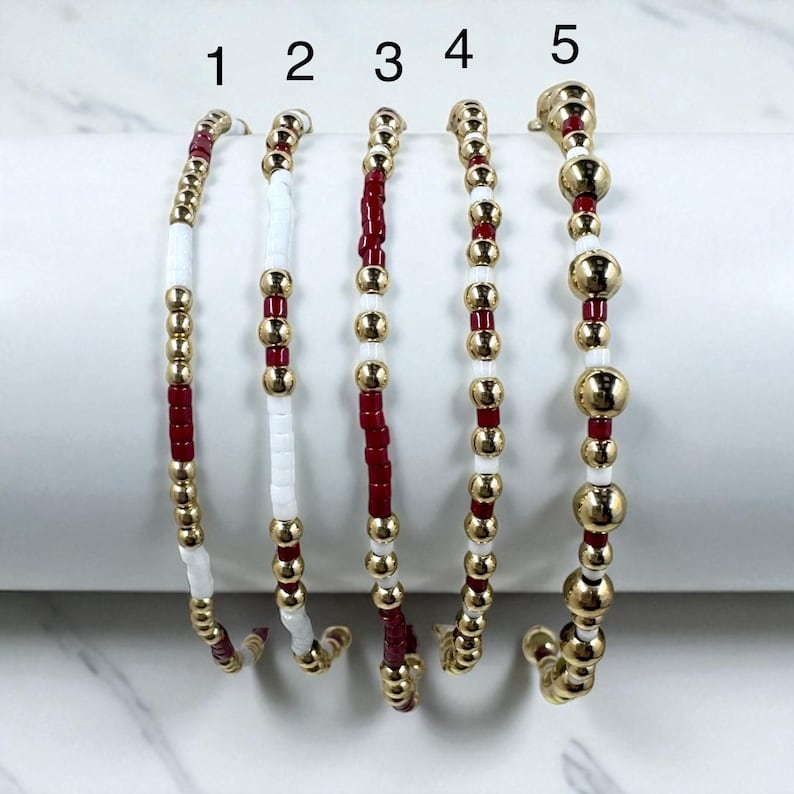 May include: Five beaded bracelets in a row. Each bracelet features a combination of gold-colored beads, white beads, and red beads. The bracelets are displayed against a white background, showcasing their colors and designs.