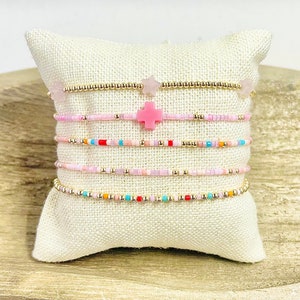 May include: A set of six beaded bracelets with pink, gold, and multi-colored beads. The bracelets are stacked on a white pillow.
